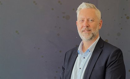 a man with a beard wearing a suit jacket stands in front of a grey wall